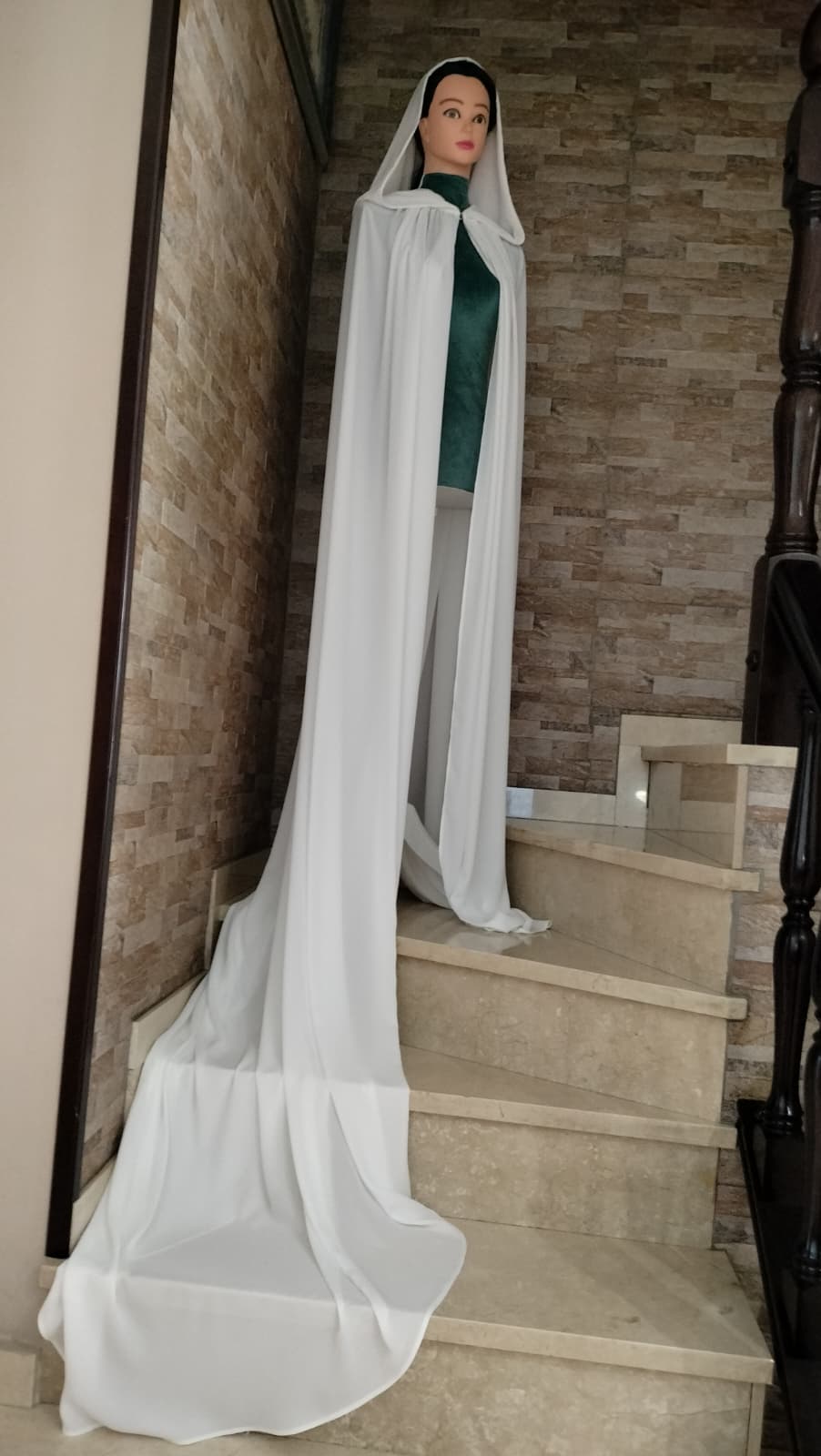 Capa de novia larga con capucha, en georgette blanco natural de Zulema Torres, extendida sobre una escalera de mármol.