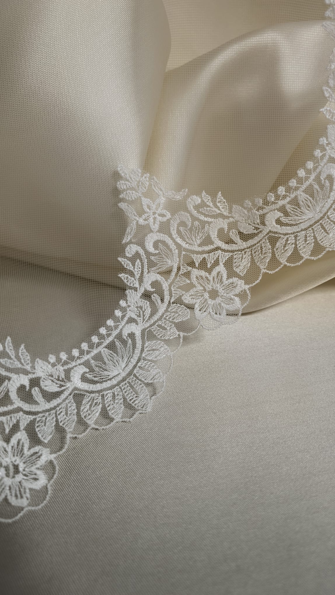 Detalle continuo del encaje de algodón calado con ondas románticas sobre un fondo satinado de color crema.