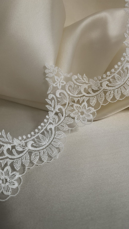 Detalle continuo del encaje de algodón calado con ondas románticas sobre un fondo satinado de color crema.