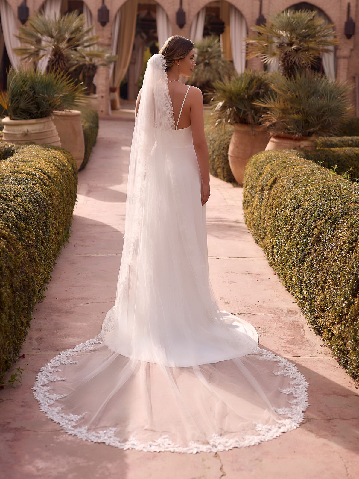 Vista posterior de una novia que lleva un vestido de novia minimalista con escote en la espalda y un velo extralargo de tul transparente con ricos apliques de encaje floral a lo largo de todo el borde. El entorno es un patio rústico con vegetación y luz natural cálida nos ofrece Zulema Torres
