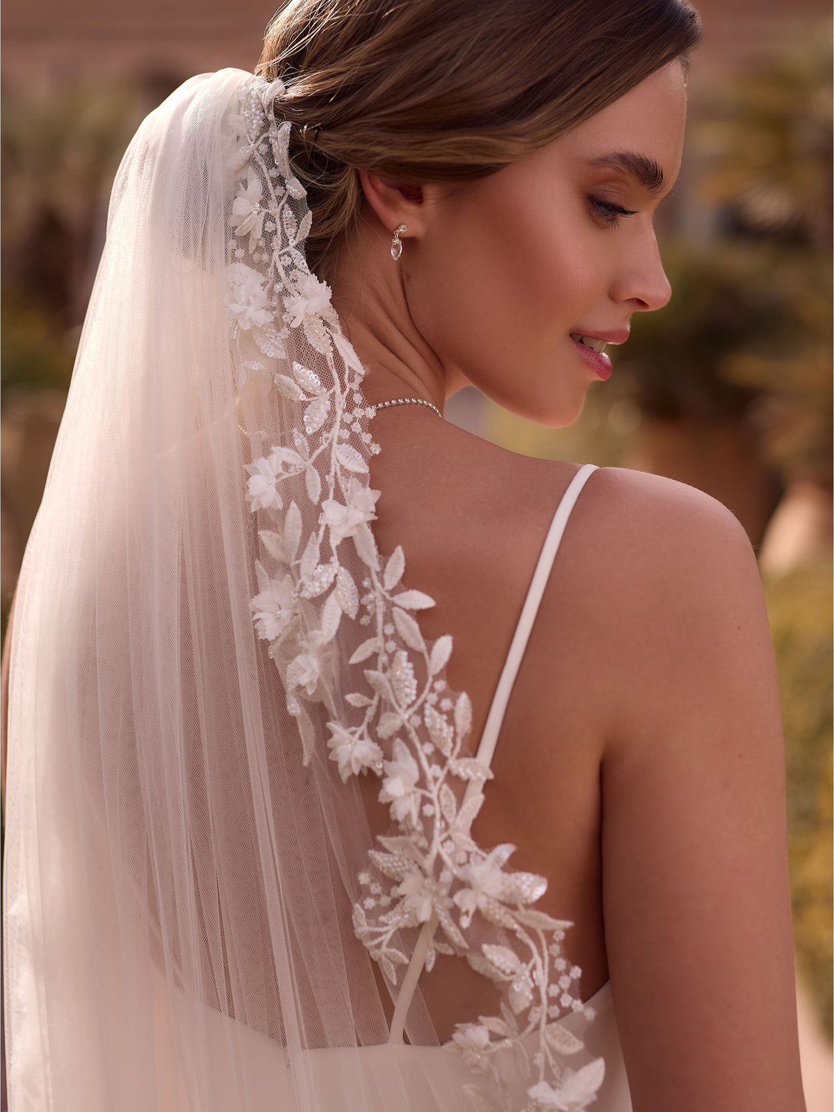 Zulema Torres nos muestra Primer plano fotográfico de perfil de una novia sonriendo suavemente. Viste un vestido blanco con tirantes finos y la espalda descubierta. Un velo de tul largo cae sobre su hombro derecho, decorado con un exquisito aplique de encaje floral 3D detallado y cuentas. Se aprecian sutilmente un pendiente colgante y un collar delicado. El fondo es un entorno luminoso con vegetación desenfocada