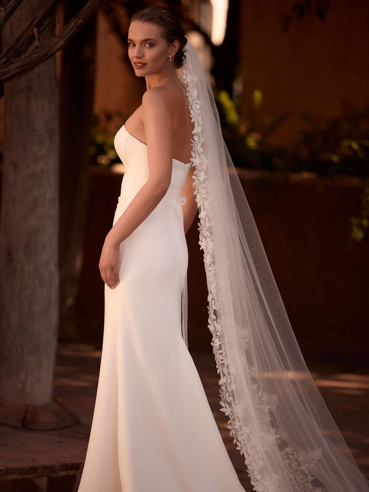 otografía de cuerpo entero y de perfil de una novia con un elegante vestido blanco ajustado de corte sirena sin tirantes, decorado con sutiles apliques de encaje en el escote y los hombros. Lleva puesto un velo de tul largo con bordes adornados con delicados motivos florales en 3D. La novia mira hacia atrás con una sonrisa suave hacia la cámara, en un entorno exterior iluminado por la luz del atardecer.Zulema Torres