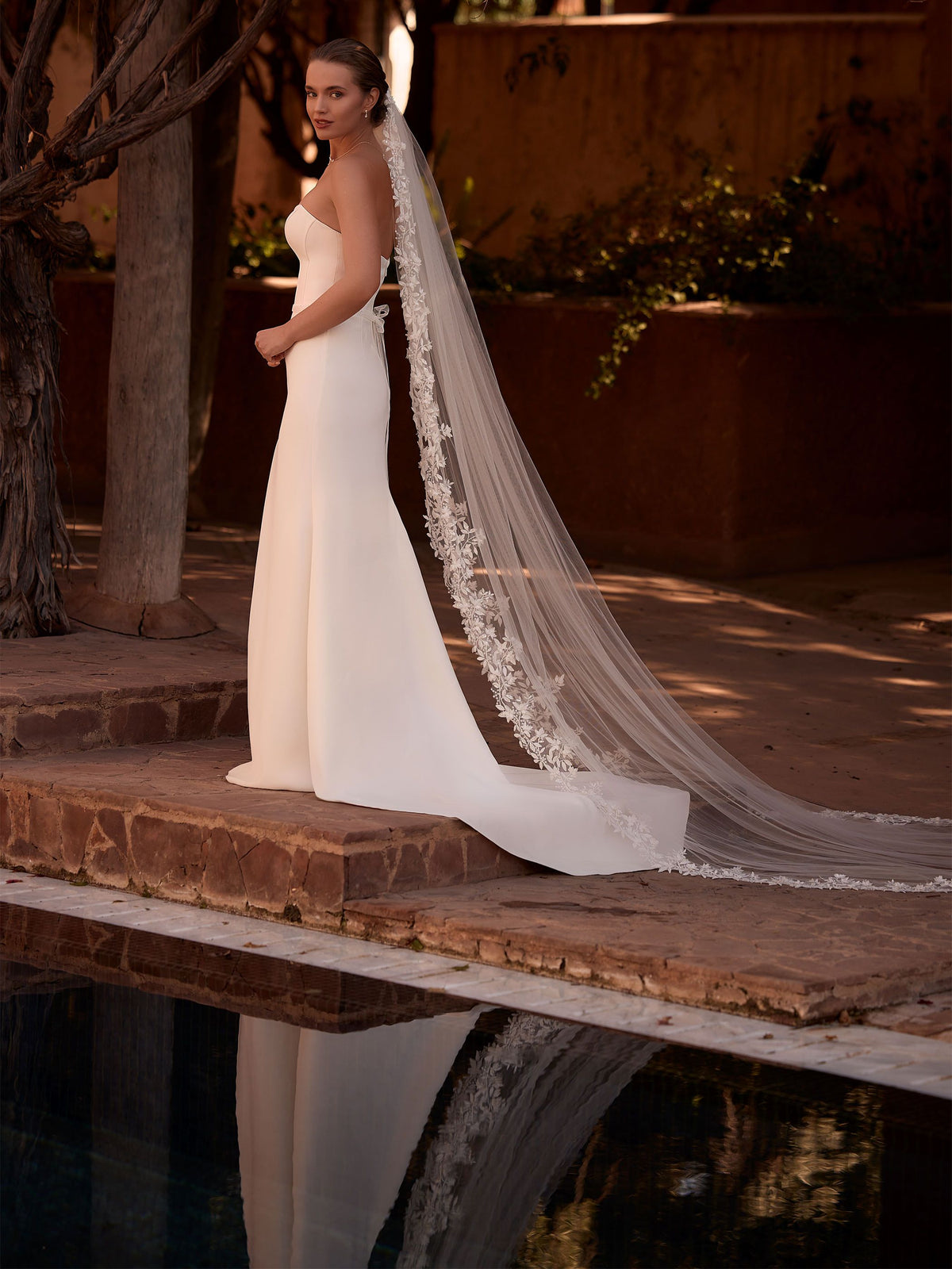 Fotografía de cuerpo entero de una novia de pie en el borde de una piscina reflectante. La novia, de perfil, lleva un elegante vestido de novia blanco, ajustado al cuerpo, con escote palabra de honor. Un velo de catedral transparente y extralargo, adornado con intrincados apliques de encaje floral a lo largo de sus bordes, se extiende dramáticamente detrás de ella sobre las baldosas de terracota y sobre la superficie del agua nos ofrece Zulema Torres