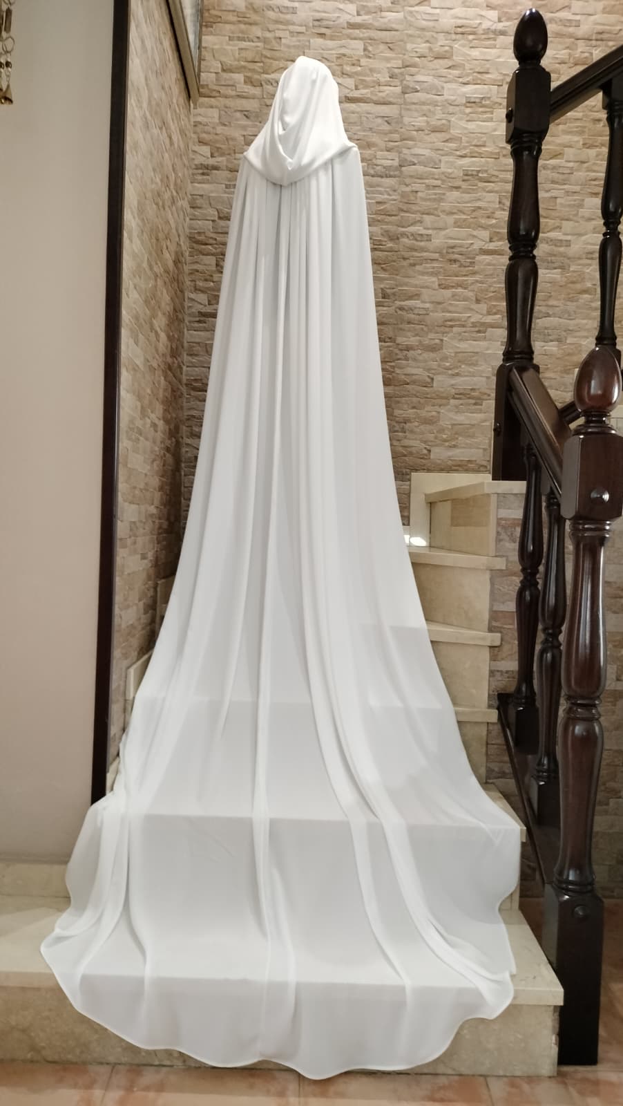 Capa de novia larga con capucha, en tejido georgette blanco natural de Zulema Torres, extendida sobre una escalera de mármol.