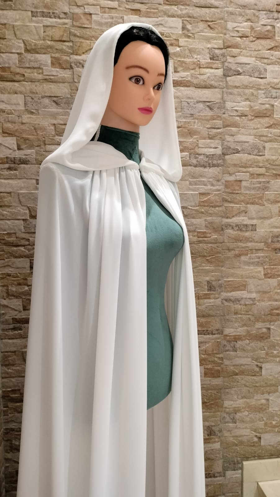 Primer plano lateral de maniquí con capa de novia blanca con capucha de georgette natural de Zulema Torres, sobre fondo de pared de piedra.