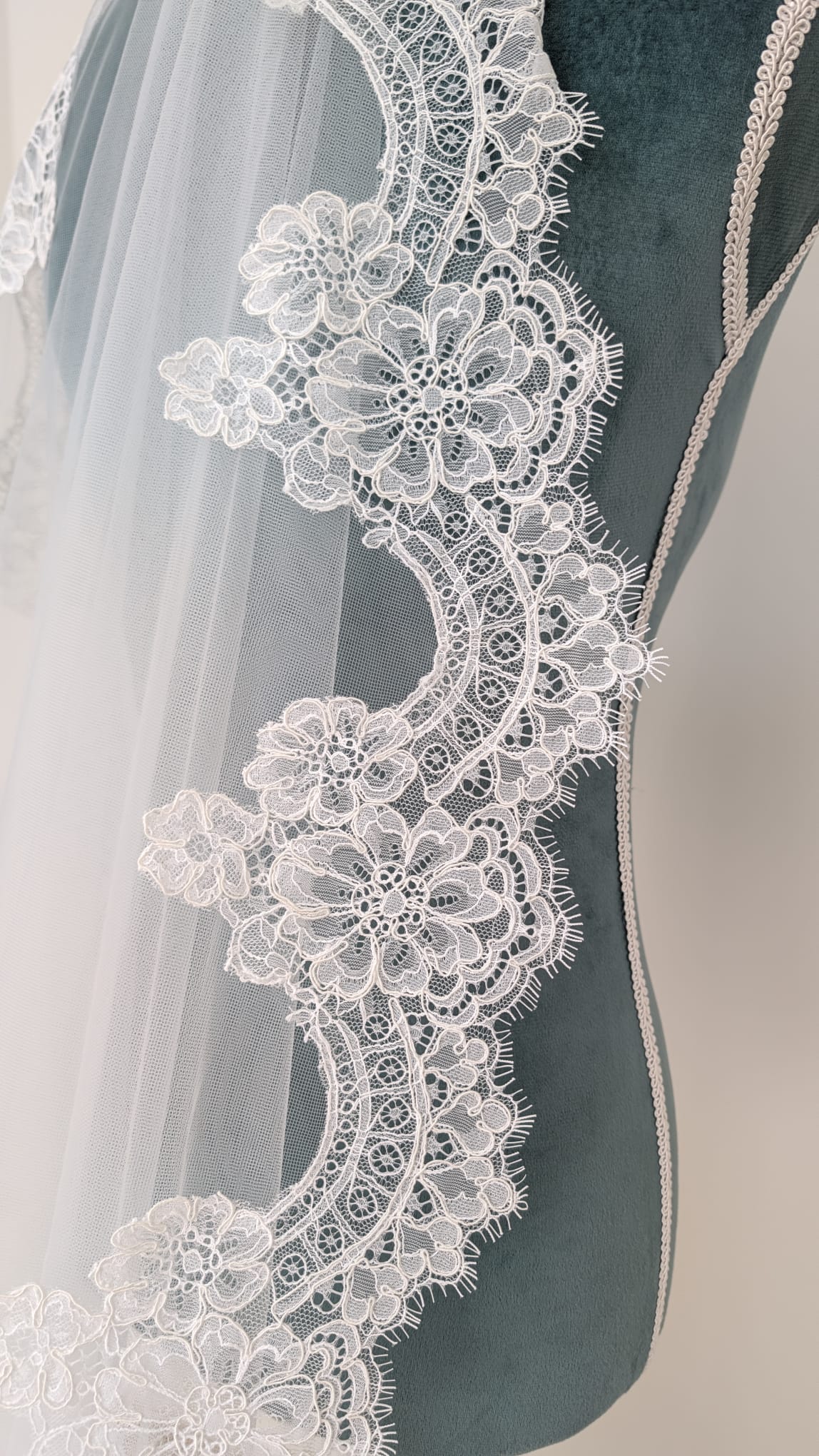 Borde de un velo de novia largo de tul sedoso con encaje floral continuo expuesto sobre un maniquí de costura verde.