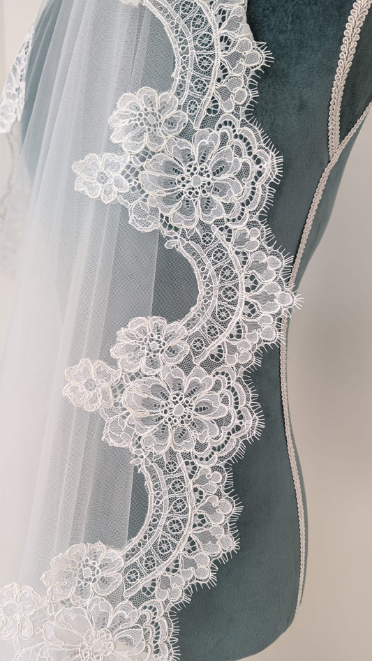 Borde de un velo de novia largo de tul sedoso con encaje floral continuo expuesto sobre un maniquí de costura verde.